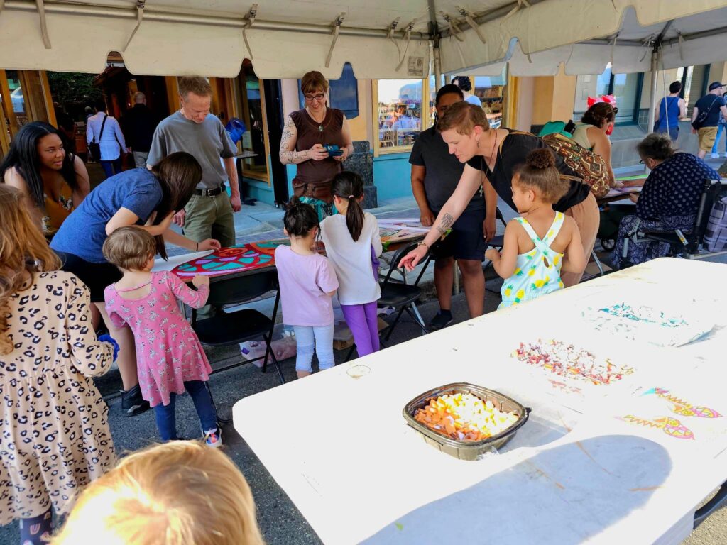 Adults and children participating in a large group mosaic project at an art festival.