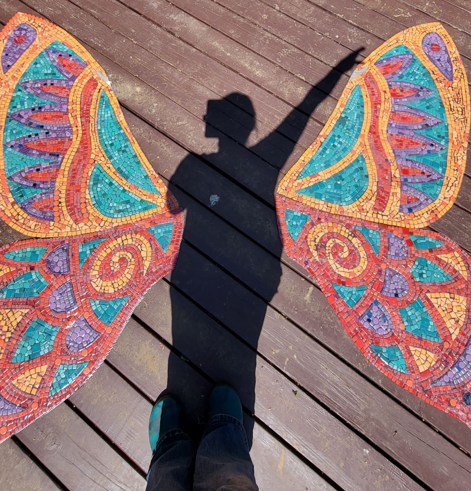 Mosaic wings in red, orange, teal and purple are laid out on a deck with a shadow standing in the center.